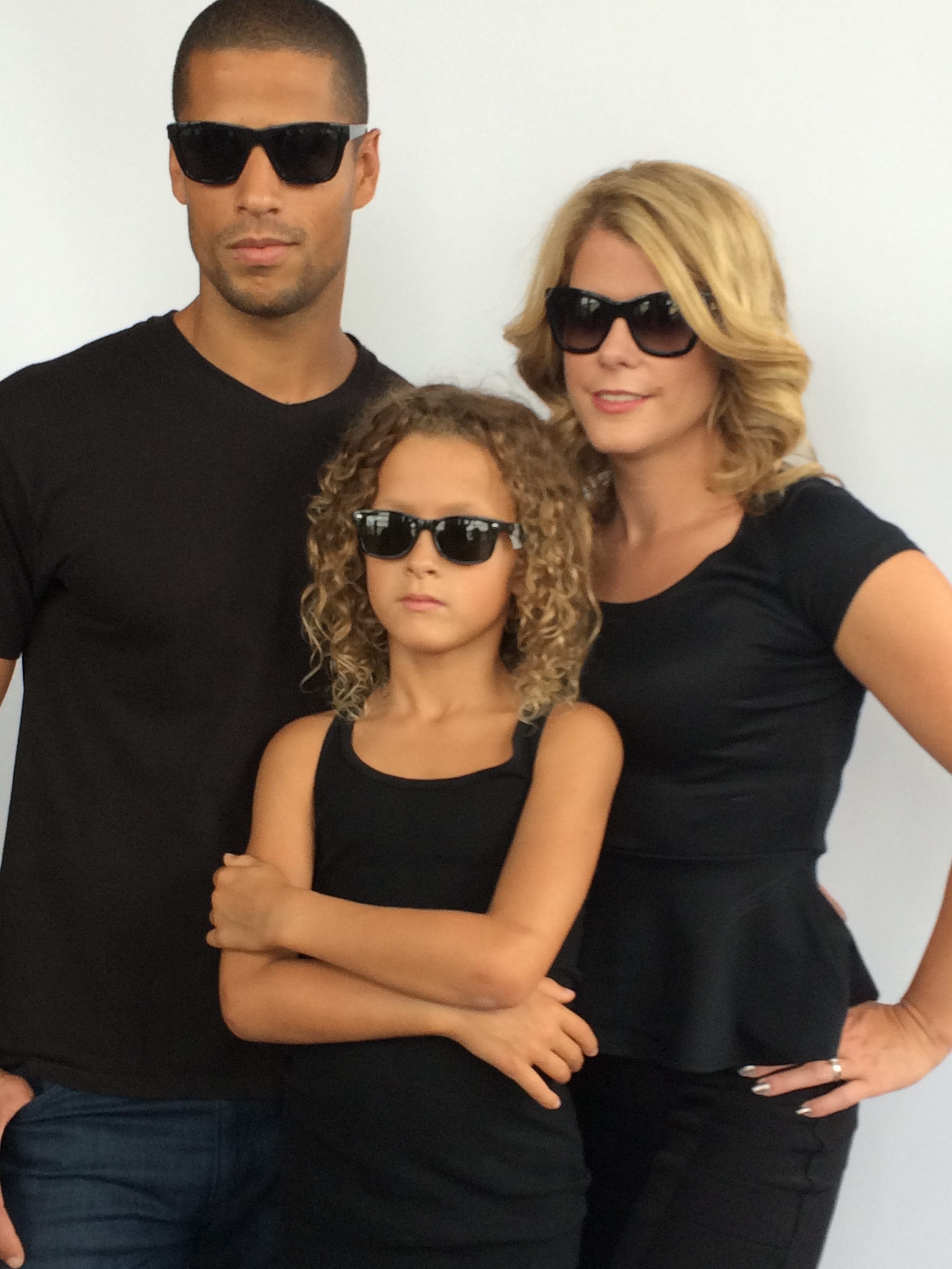 Black male dad, blonde female mom, curly haired boy posing wearing black plastic sunglass frames with dark lenses. Blonde female wearing turquoise plastic designer frame Maple Grove Eye Doctors at Pearle Vision