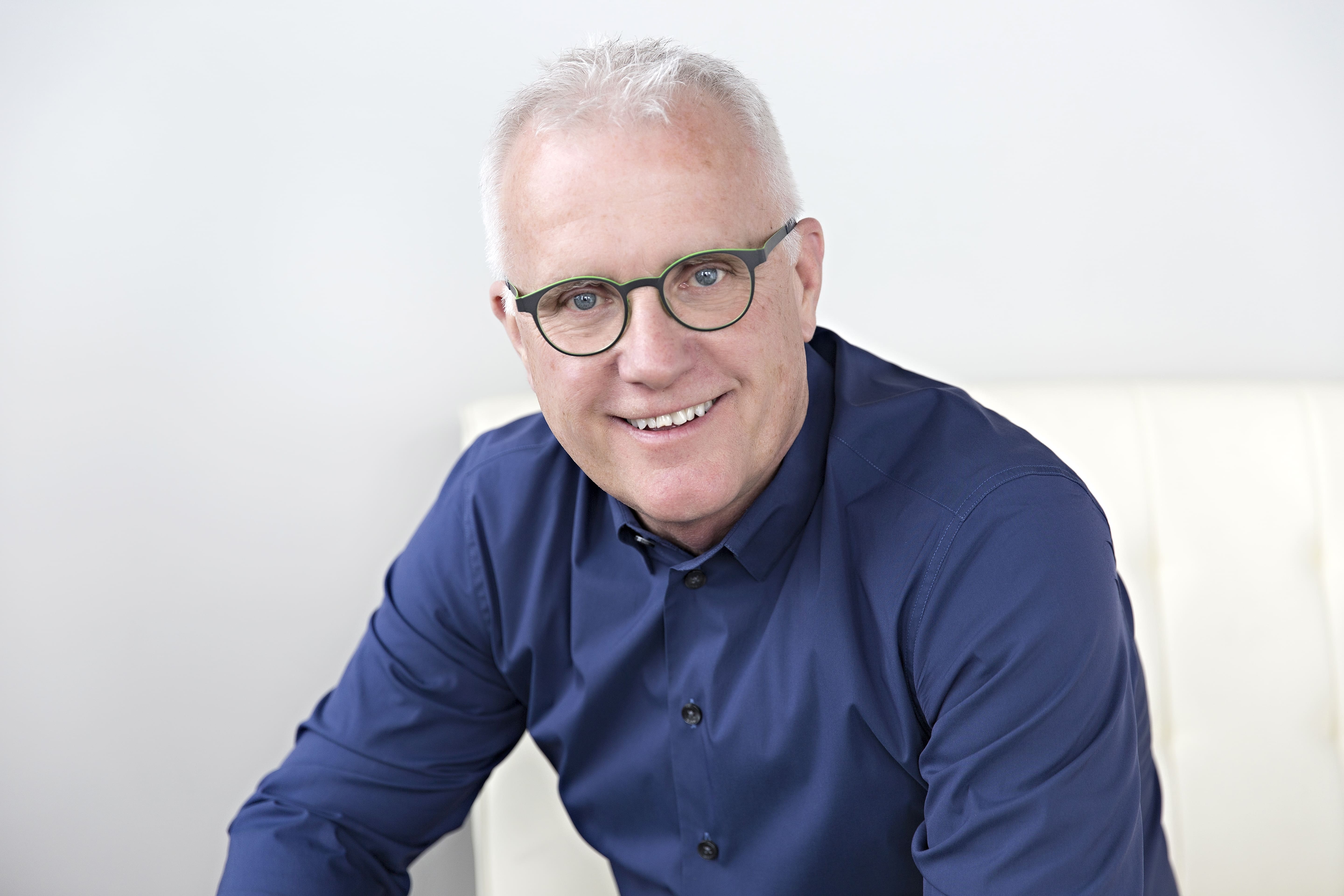 Grey haired male in blue shirt wearing green round frames while smiling and posing Maple Grove Eye Doctors at Pearle Vision