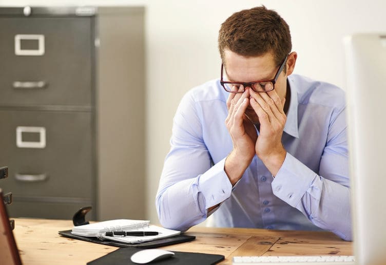 Man in dress shirt at desk rubbing his eyes under his glasses while sitting at a desk. Maple Grove Eye Doctors at Pearle Vision