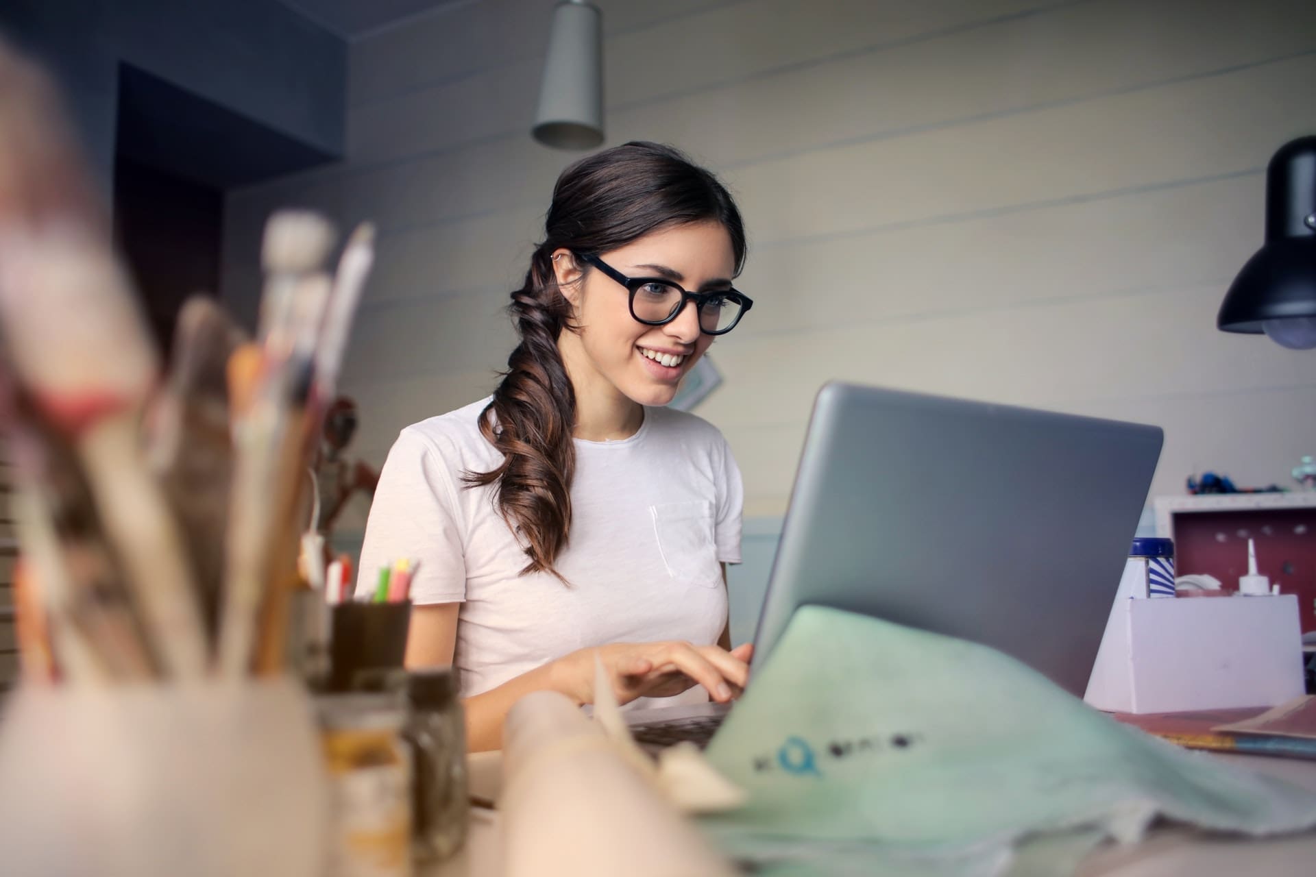 Young dark haired woman with long side ponytail smiling while working on laptop wearing black plastic designer frame Maple Grove Eye Doctors at Pearle Vision
