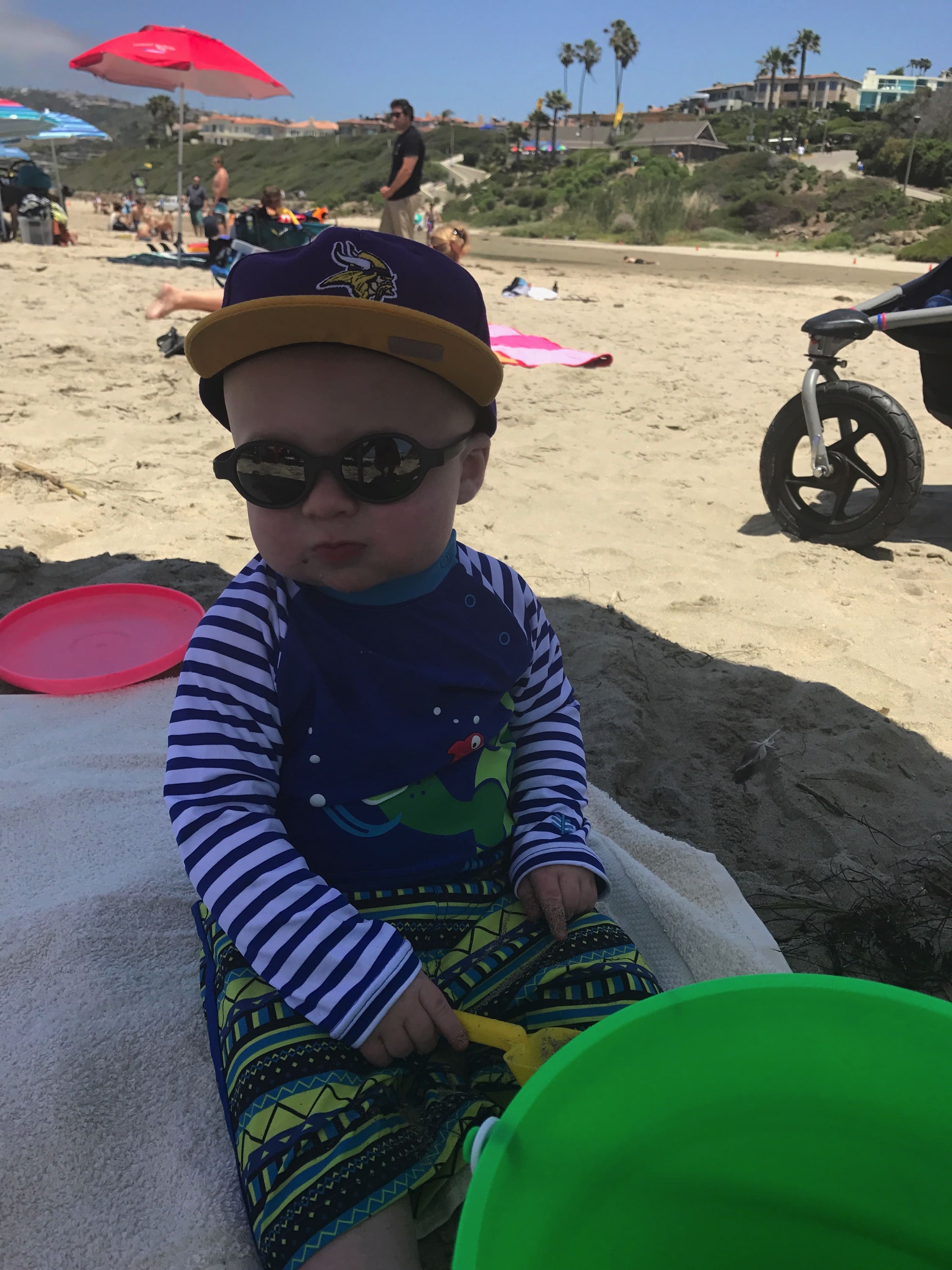 Baby with hat and sunglasses siting at the beach Maple Grove Eye Doctors at Pearle Vision