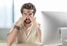 Brown curly haired man rubbing eyes with glasses on forehead in front of a laptopMaple Grove Eye Doctors